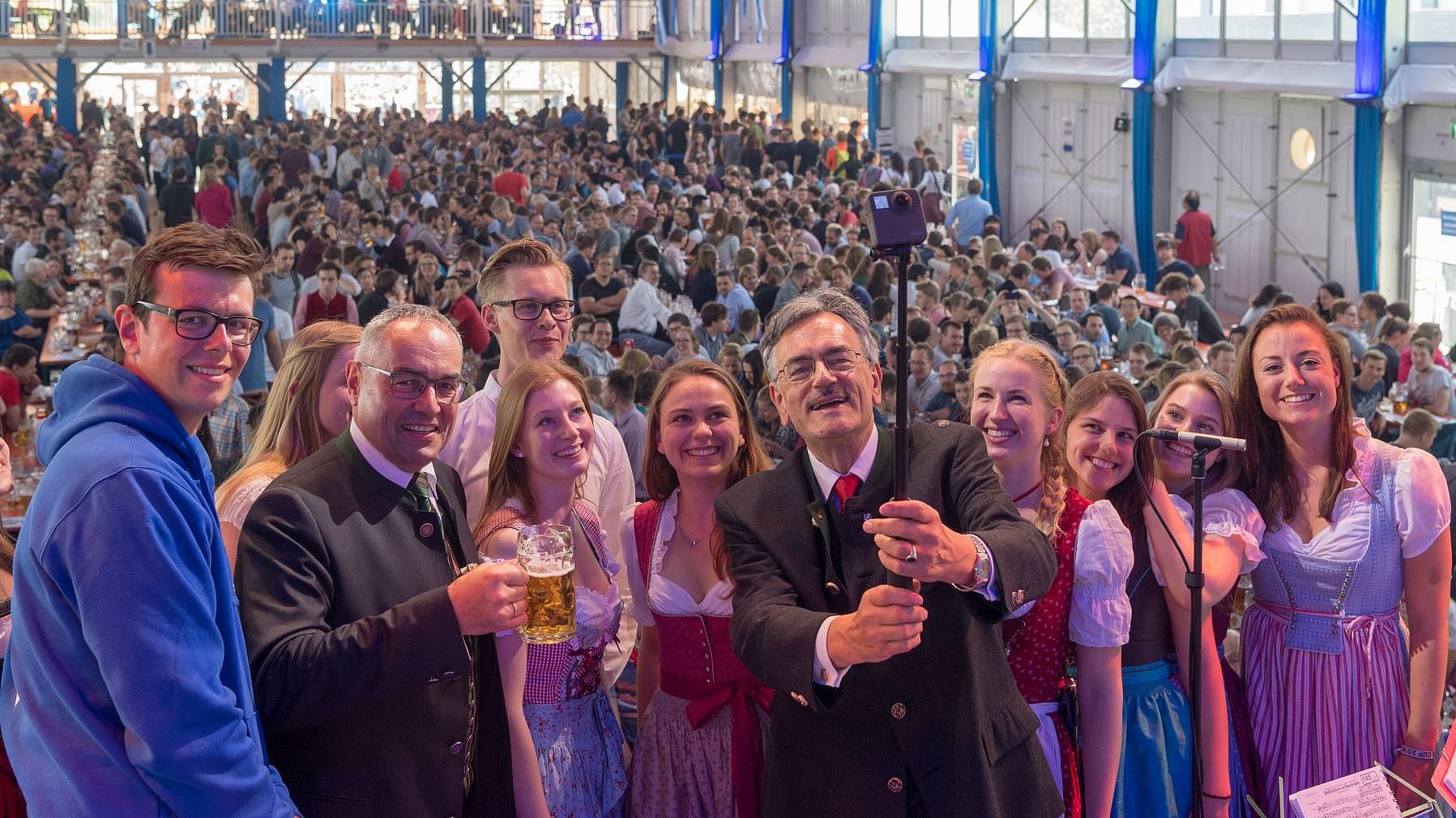 Präsident Emeritus Wolfgang A. Herrmann, im Jubiläumsjahr 2018 amtierender TUM-Präsident, und Kanzler Albert Berger (r.) mit Studierenden mit einem Selfiestick im Festzelts des Mitarbeiterfests im Jubiläumsjahr 2018, hinter ihnen zahlreiche Gäste an Bierbänken. 