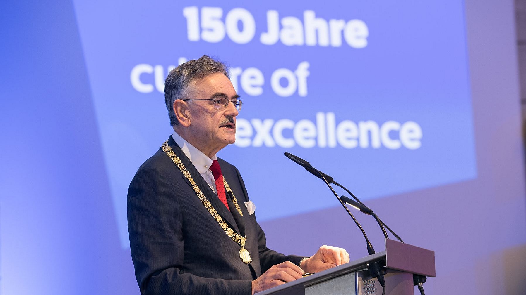 Wolfgang A. Herrmann spricht in ein Mikrofon. Hinter ihm ist eine Präsentatipn mit dem Schriftzug „150 Jahre culture of excellence“. 