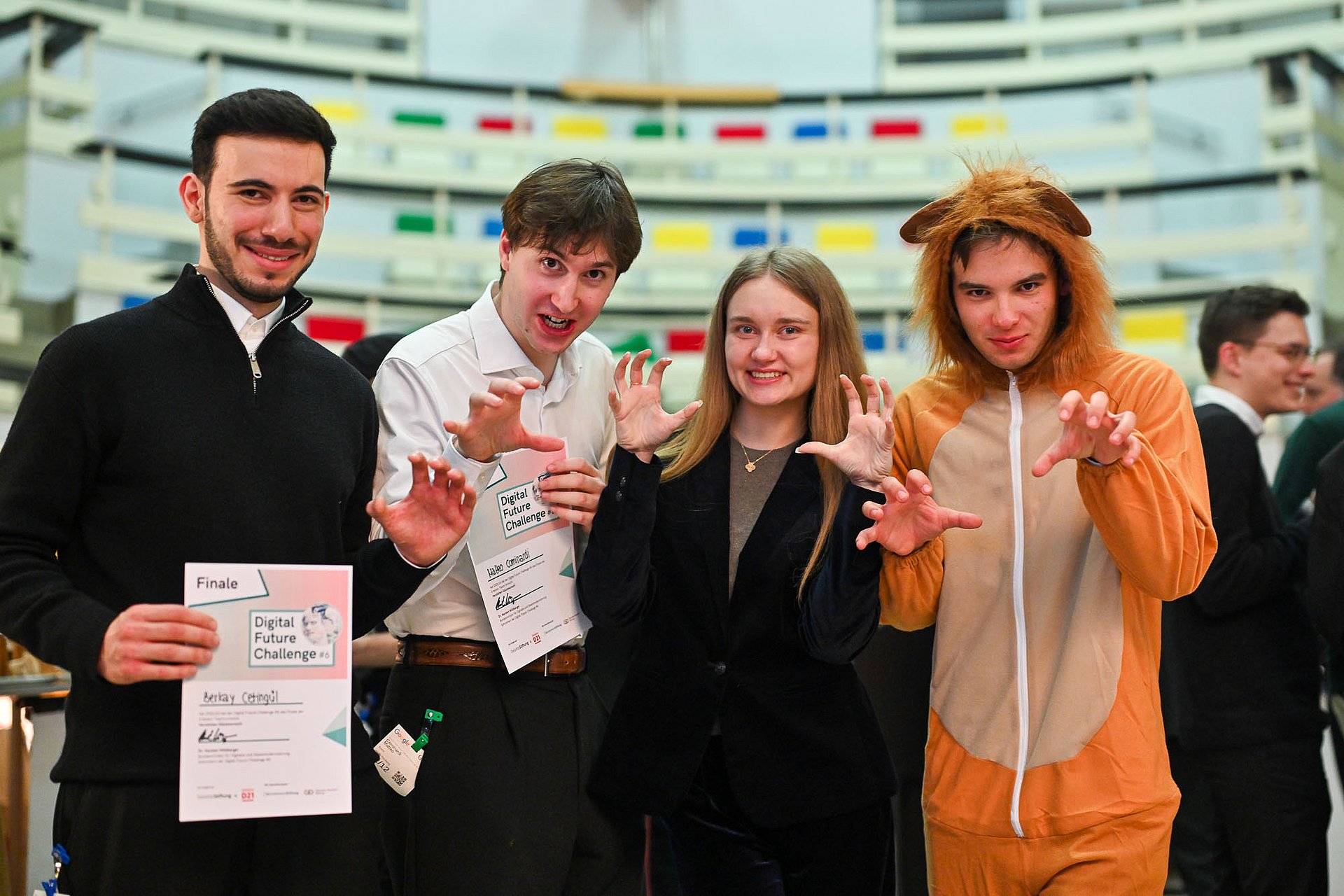 Team “Lioness & the Three Lions” at the final of the Digital Future Challenge: The four team members pose for the photo. Two people hold certificates from the Digital Future Challenge. All four raise their hands in a playful lion gesture. One person is wearing a lion costume.