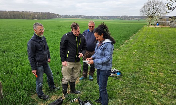 Eine Frau und drei Männer stehen auf einem Acker. Die Frau zeigt den Männern ein Gerät in ihrer Hand. Es handelt sich um das Gerät, dass das NutriSen-Team zur smarten Düngung entwickelt hat.