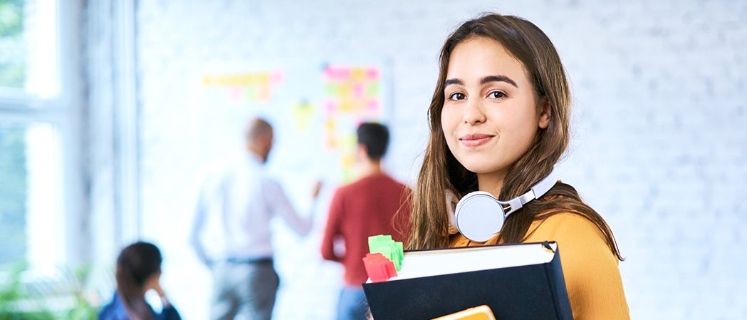 In die Kamera blickende Studentin, im Hintergrund ein heller, moderner Seminarraum