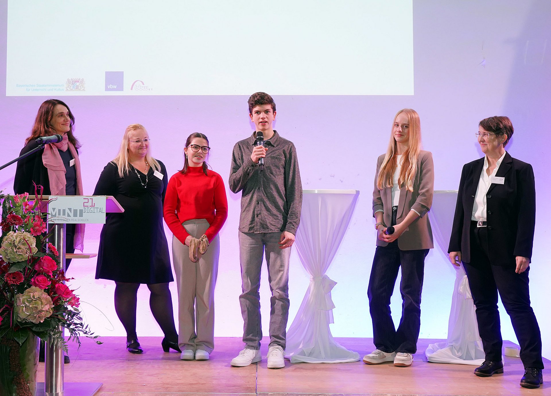 Eine Gruppe von sechs Personen, davon drei erwachsene Frauen, zwei Schülerinnen und ein Schüler, auf der Bühne bei der Verleihung des MINT21-Digital-Preises an der Staatlichen Realschule in Herrsching. Der Schüler steht mittig platziert und spricht in ein Handmikrofon.
