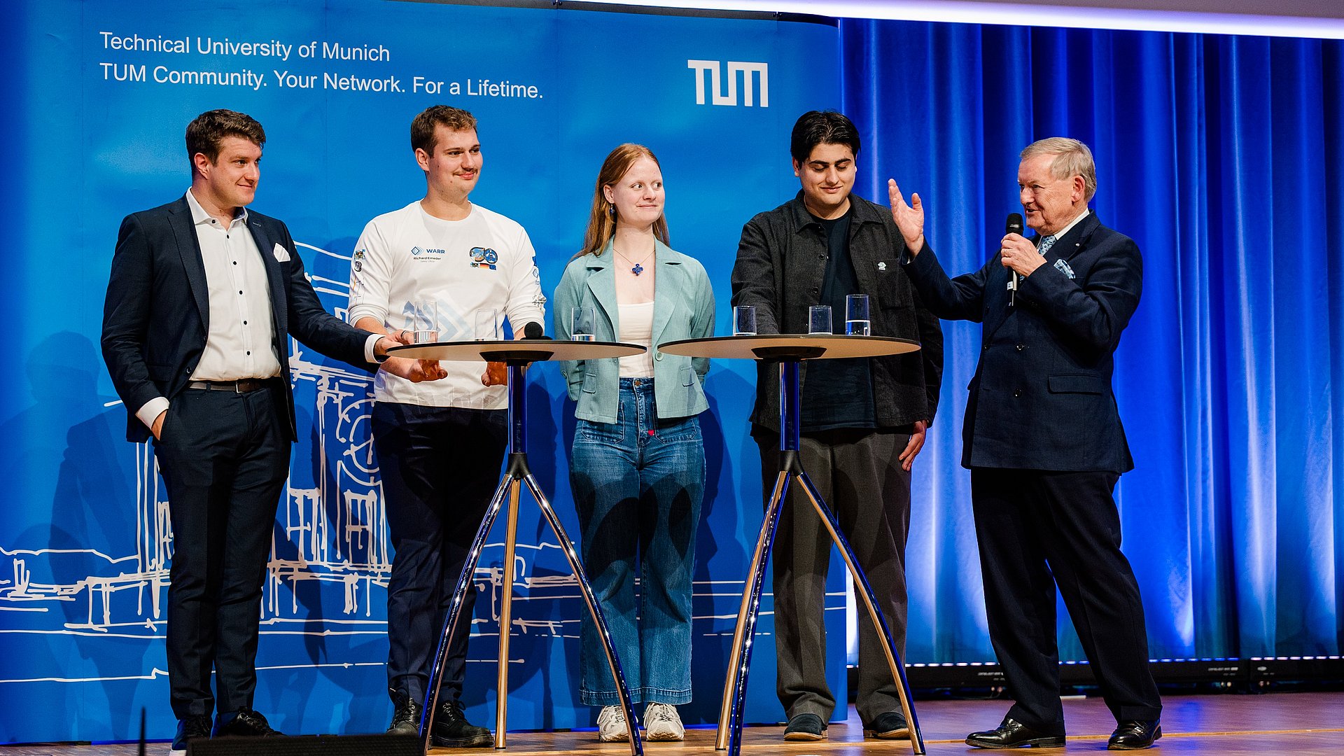 Fünf Personen auf einer Bühne vor blauem Hintergrund an zwei Stehtischen stehend, von links nach rechts: Josef Fleischmann, Richard Emeder, Mareike Ufer, Ceyhun Aydin und Robert Schmucker; Herr Schmucker spricht, den anderen vieren zugewandt, gerade in ein Handmikrofon und gestikuliert mit der rechten Hand.