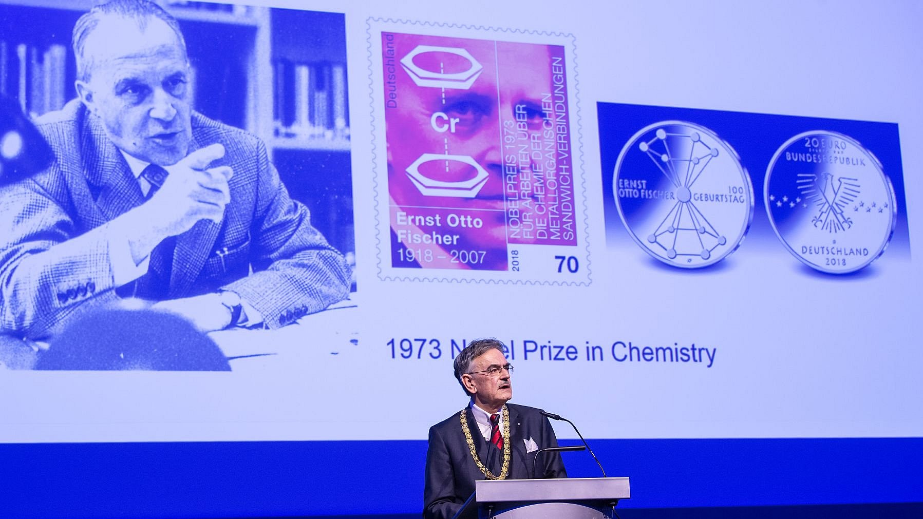 Präsident Emeritus Wolfgang A. Herrmann, im Jubiläumsjahr 2018 amtierender TUM-Präsident, hält eine Rede bei der akademischen Jahresfeier „Dies Academicus“ im Jahr 2018. Im Hintergrund ist eine Präsentation mit Fotos des Nobelpreisträgers Ernst Otto Fischer zu sehen. 