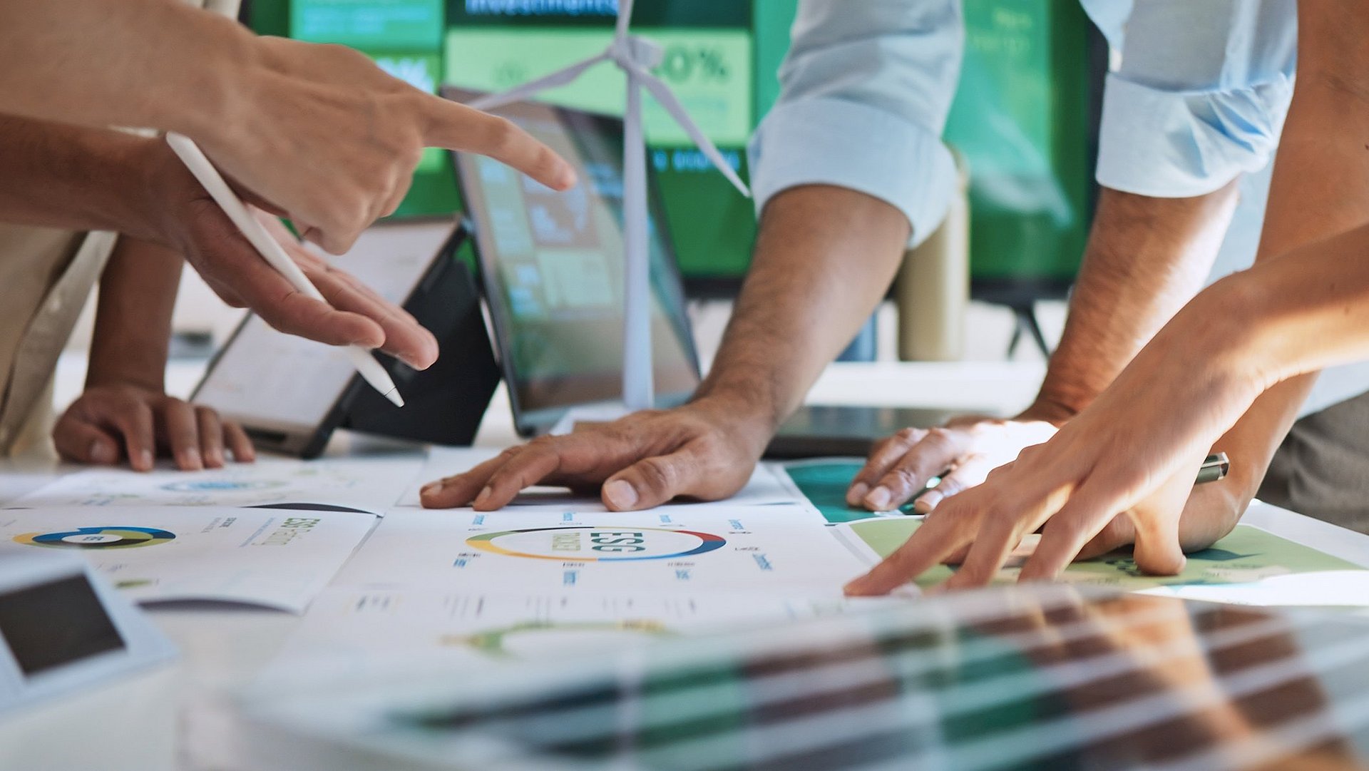 Es liegen Blätter mit verschiedenen Diagrammen auf einem Tisch. Darauf stützen sich mehrere Personen, die über die Diagramme diskutieren. Zu sehen sind allerdings nur deren Hände.