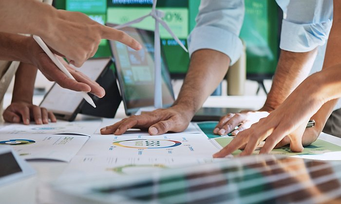 Es liegen Blätter mit verschiedenen Diagrammen auf einem Tisch. Darauf stützen sich mehrere Personen, die über die Diagramme diskutieren. Zu sehen sind allerdings nur deren Hände.