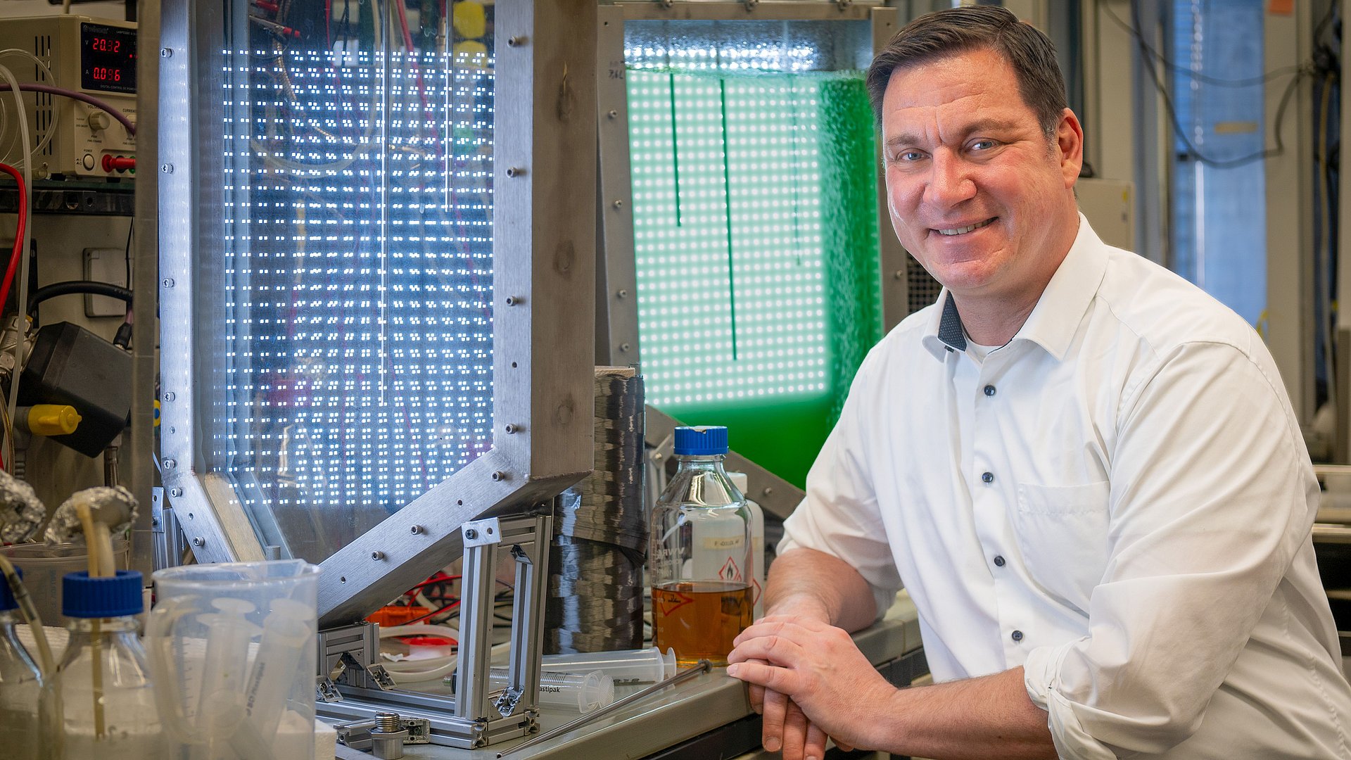 A man sits in the labortary. On the work desk, there is a spool with grey-silver material wrapped around it. In the back, there is a green-lit tank visible.