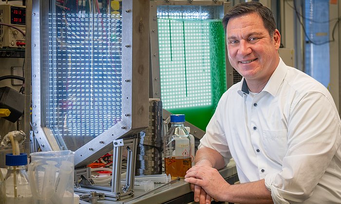 Ein Mann sitzt auf einem Laborstuhl vor einer Arbeitsplatte. Vor ihm steht eine Spule mit grau-silbernem Material. Außerdem ist ein grün leuchtender Tank zu sehen.