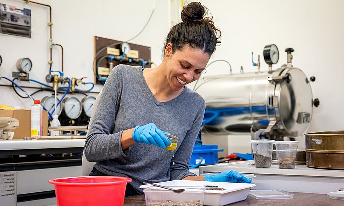 Eine Frau sitzt am Tisch mit einer kleinen Schale in der Hand. Auf dem Tisch stehen mehrere Behältnisse mit Erde.