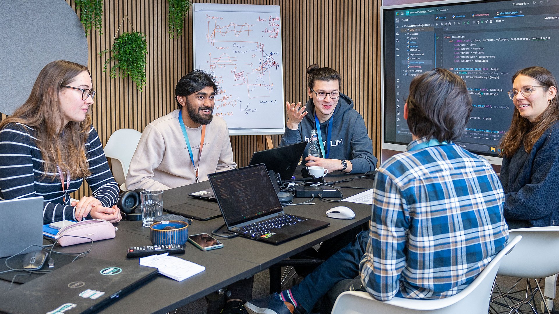 Five students sit around a table and discuss. There is a laptop in front of two students. Source code can be seen both on the laptops and on the screen in the background.