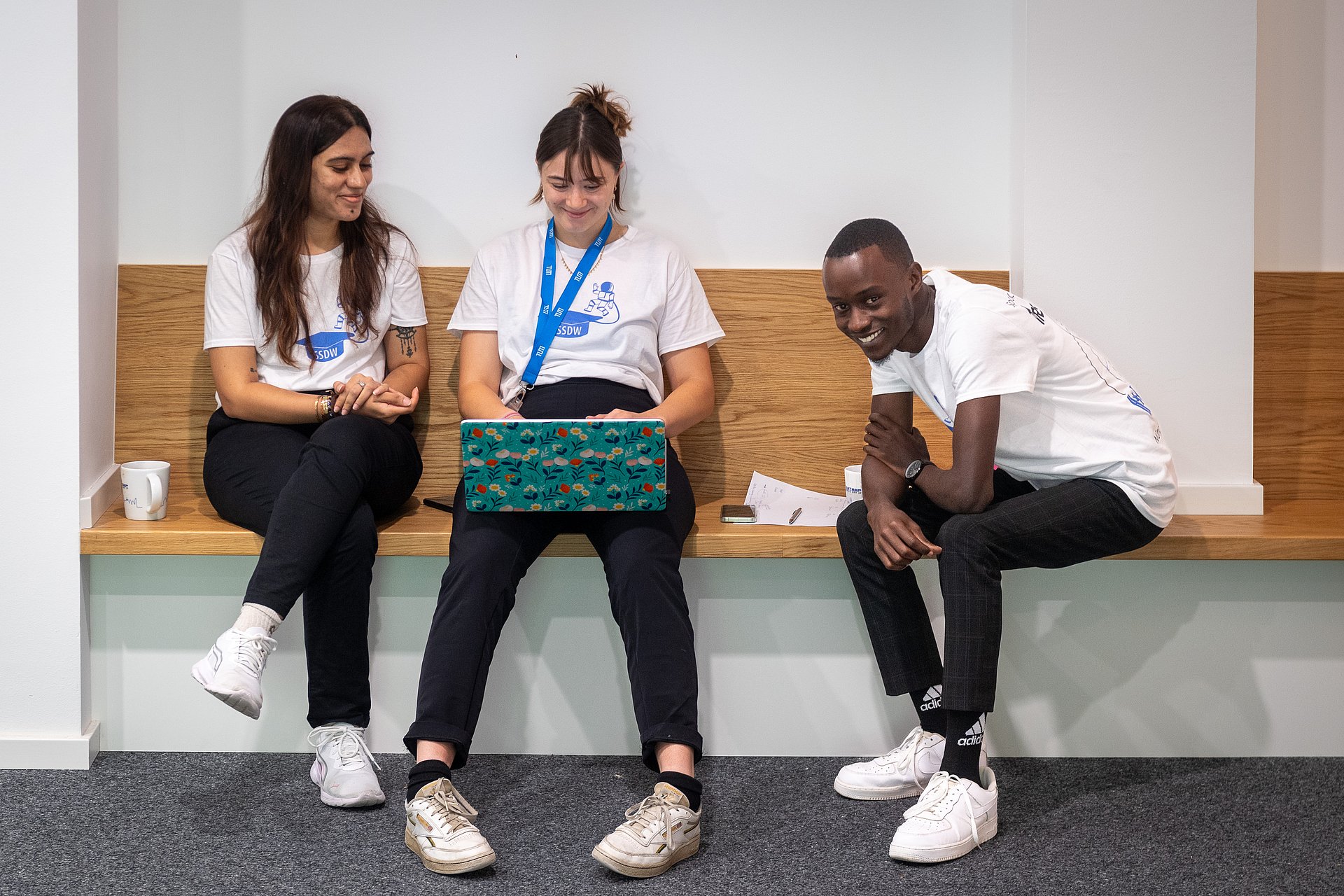 Drei Teilnehmende sitzen freudestrahlend in einer ruhigen Ecke und arbeiten am Laptop