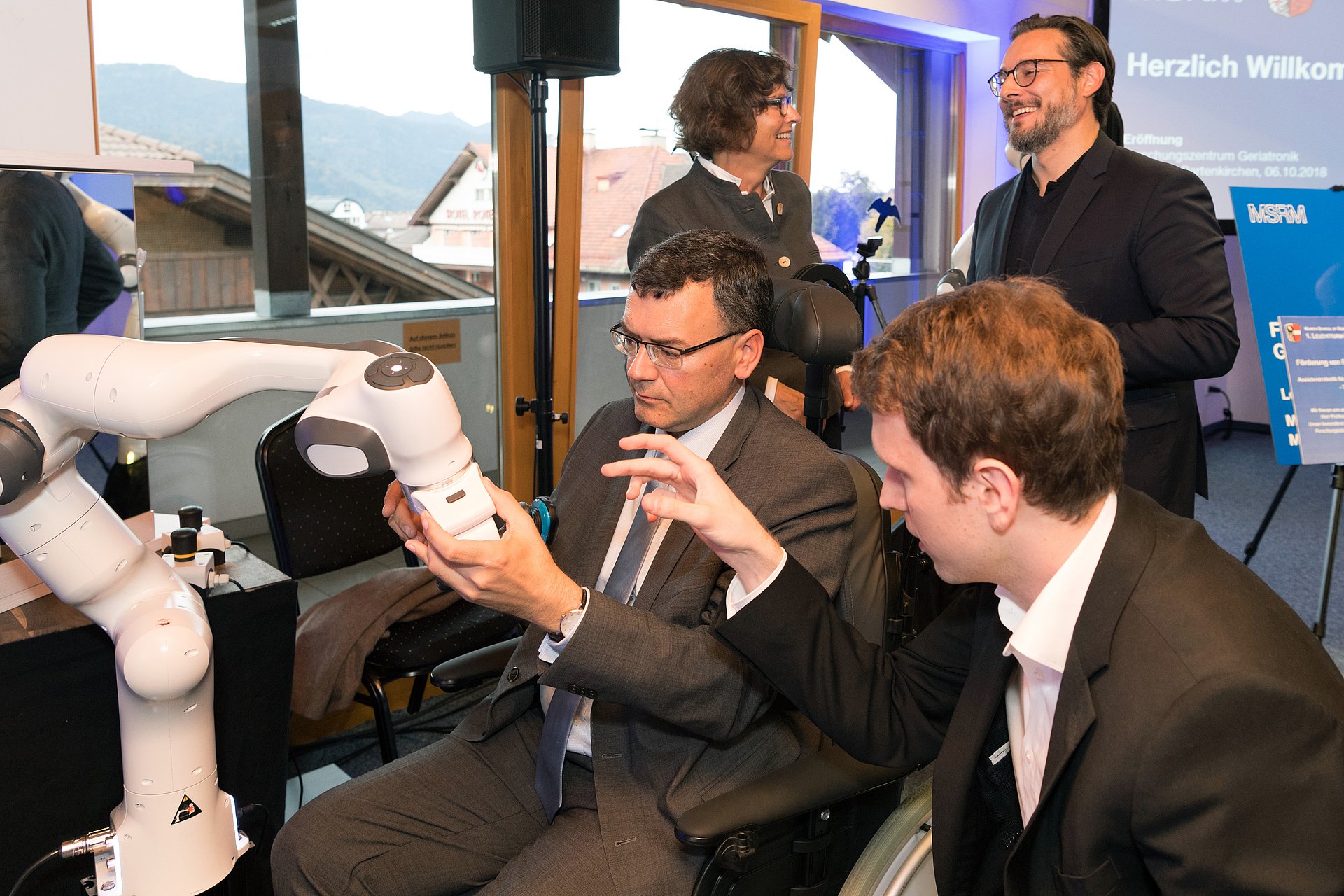 Bavarian State Mnister Dr. Florian Hermann tests a shaving robot. In the background, Dr. Sigrid Meierhofer, mayor of Garmisch-Partenkirchen and Professor Sami Haddadin are talking.