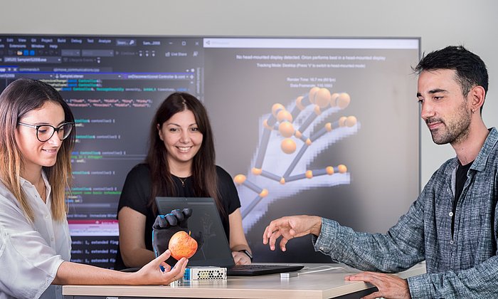 Drei Personen, die um einen Tisch stehen und eine Handprothese testen. Die Frau links im Bild hält einen Apfel in der Hand, nach dem die Handprothese greift.