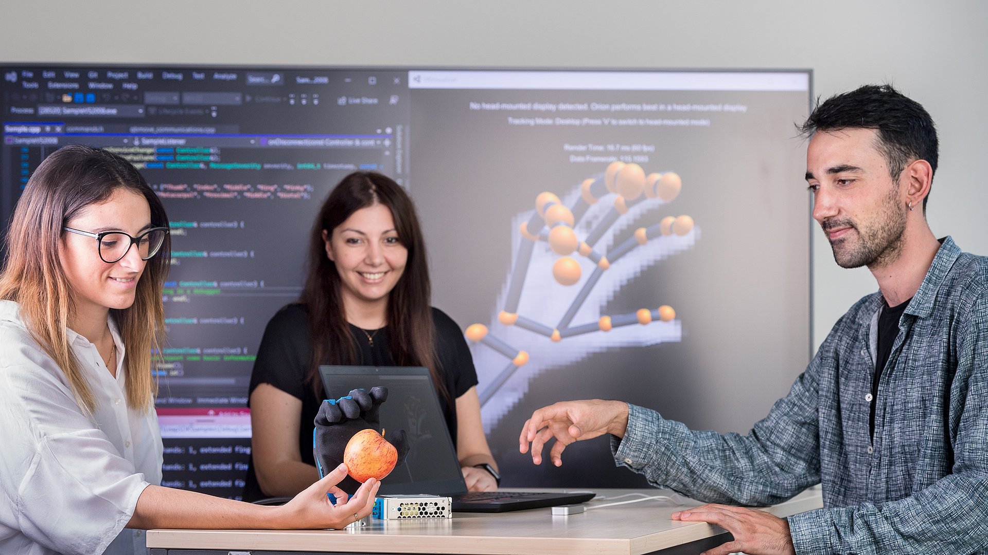 Drei Personen, die um einen Tisch stehen und eine Handprothese testen. Die Frau links im Bild hält einen Apfel in der Hand, nach dem die Handprothese greift.