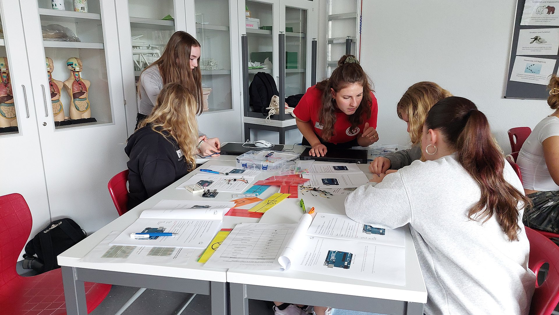 Anna Eckl, eine der Referentinnen des MINT-Impulse-Programms, dem Betrachter zugewandt über einen Tisch gebeugt, an dem vier Schülerinnen sitzen und auf dem Unterlagen und Kabel verteilt sind, mit deren Hilfe sie Sensoren bauen können.