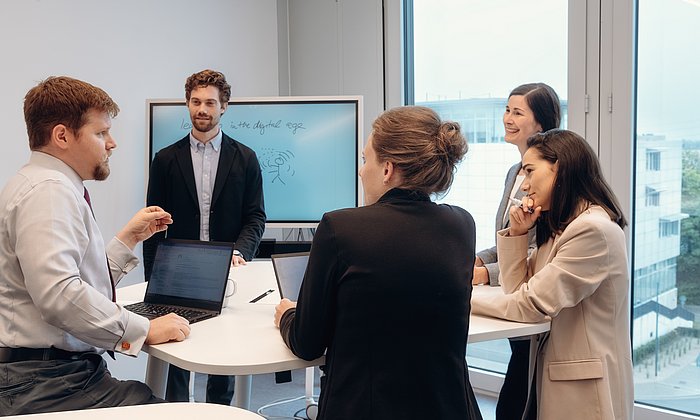 Mehrere Personen stehen in einem Seminarraum am Tisch und nehmen an einem Workshop teil.