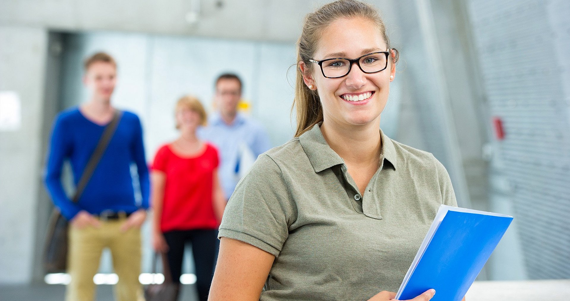Eine Studentin steht lächelnd im Raum und hält eine blaue Mappe in der Hand, während im Hintergrund drei weitere Studierende unscharf zu erkennen sind.