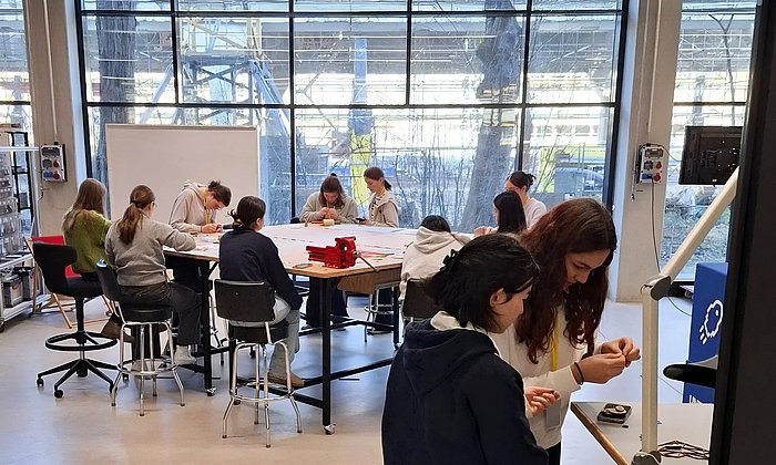 Ein heller Raum mit großen Glasfenstern, durch die eine Außenkonstruktion sichtbar ist. Im Hintergrund sitzen mehrere Schülerinnen um einen großen Tisch und arbeiten gemeinsam an Projekten oder Zeichnungen. Im Vordergrund stehen zwei Mädchen an einem Tischwagen und beschäftigen sich mit Materialien. Der Raum wirkt wie ein kreatives Atelier oder Workshop-Bereich mit offenen Regalen, einem Whiteboard und moderner Einrichtung.