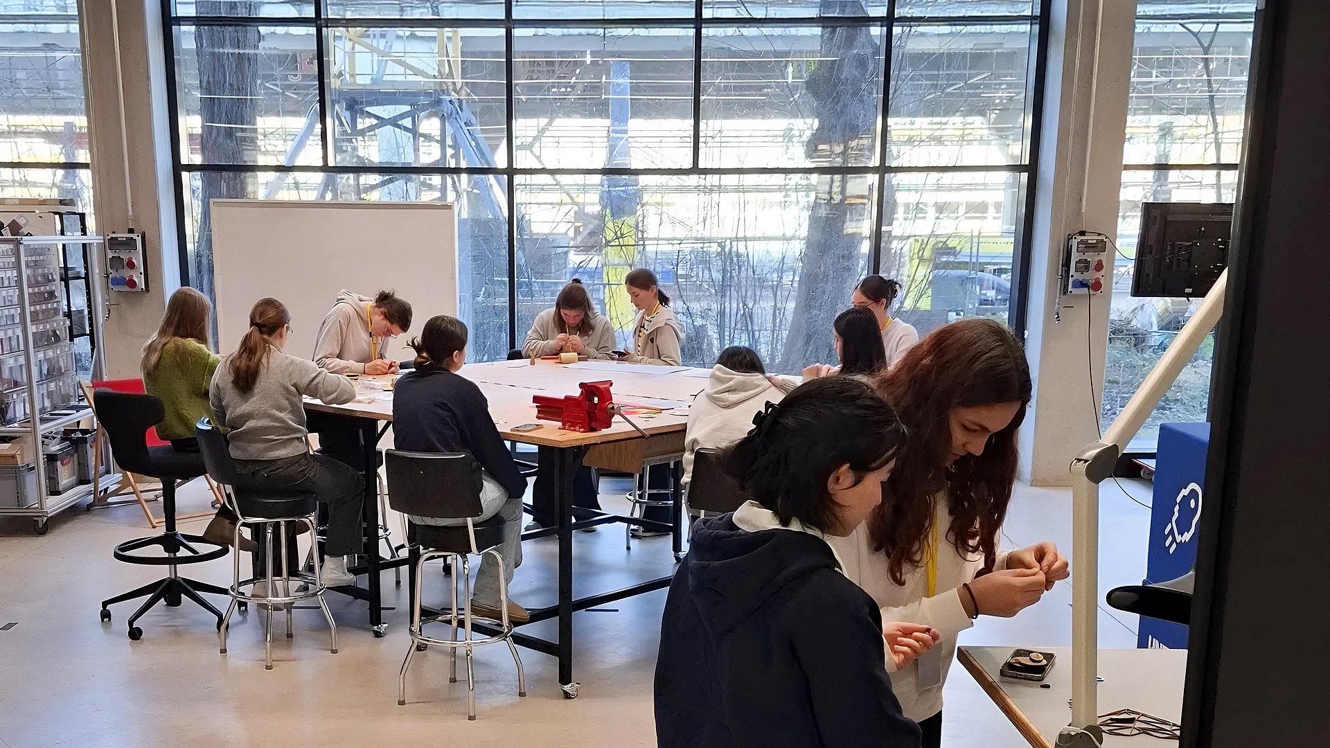 Ein heller Raum mit großen Glasfenstern, durch die eine Außenkonstruktion sichtbar ist. Im Hintergrund sitzen mehrere Schülerinnen um einen großen Tisch und arbeiten gemeinsam an Projekten oder Zeichnungen. Im Vordergrund stehen zwei Mädchen an einem Tischwagen und beschäftigen sich mit Materialien. Der Raum wirkt wie ein kreatives Atelier oder Workshop-Bereich mit offenen Regalen, einem Whiteboard und moderner Einrichtung.