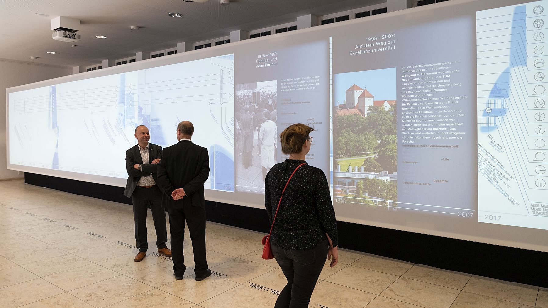 Installation der Ausstellung „Zeitlupe“ in der Immatrikulationshalle der TUM und einer Besucherin und zwei Besuchern.