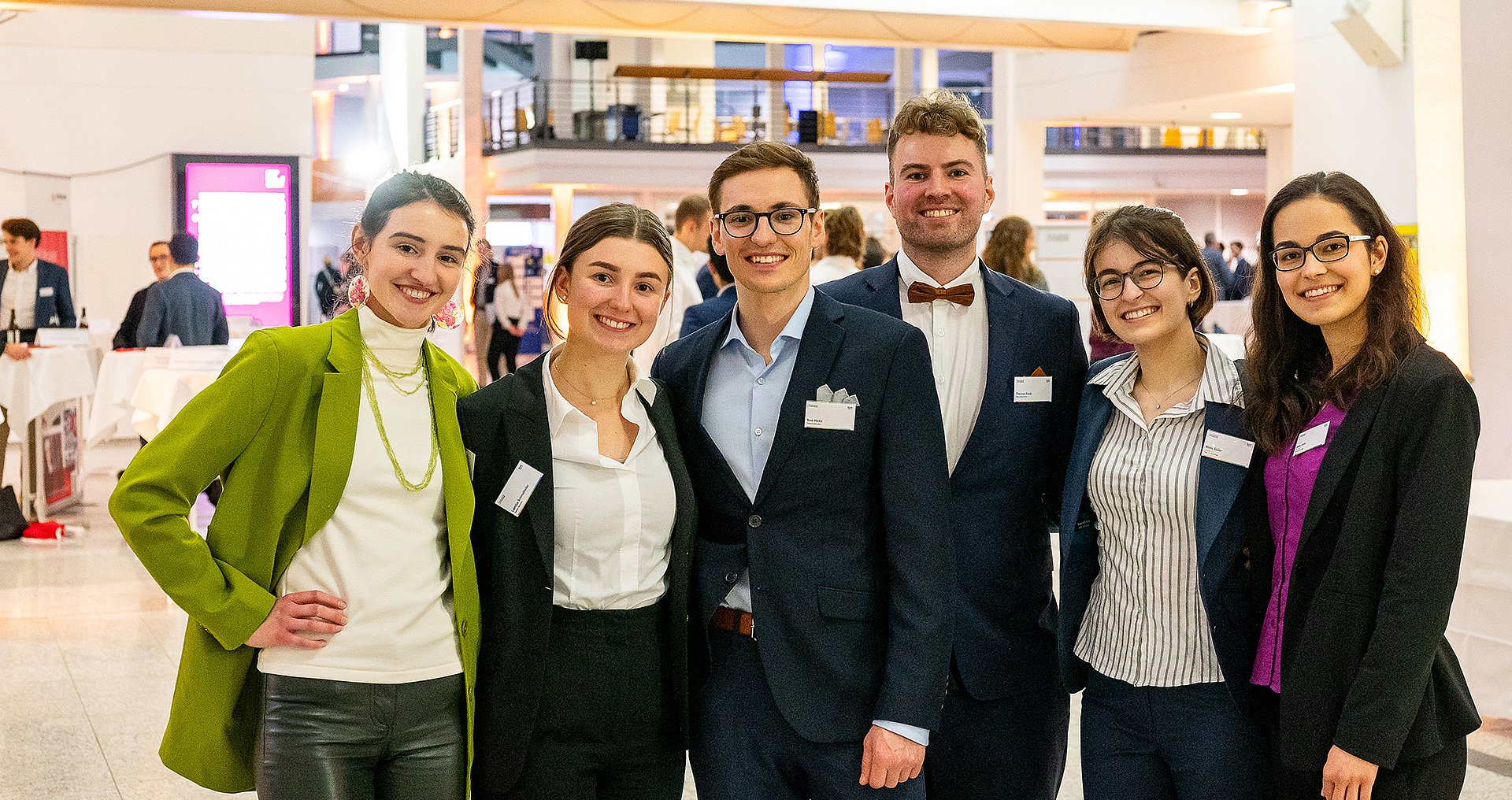 eine Gruppe von Studierenden zwei Männer und 4 Frauen, die alle Businesskleidung tragen, stehen eng beieinander und schauen lächelnd in die Kamera. 