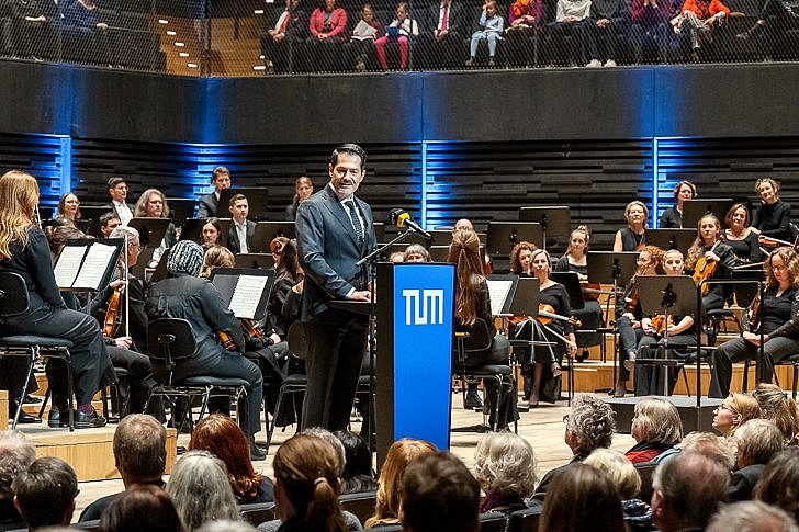 TUM President Prof. Hofmann stands on the stage and gives a welcoming speech. Behind him are the musicians of the SEM Orchestra