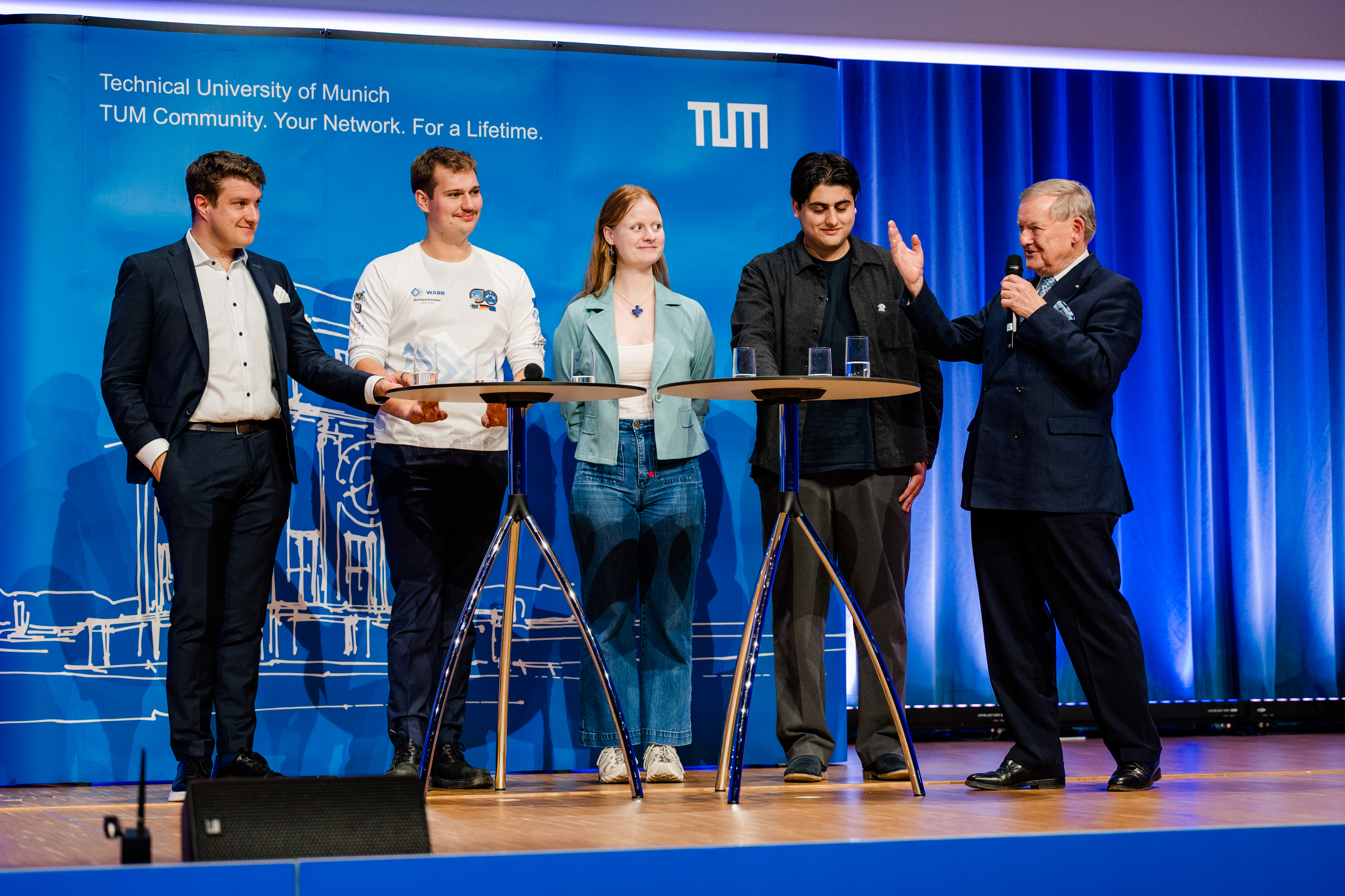 Fünf Personen auf einer Bühne vor blauem Hintergrund an zwei Stehtischen stehend, von links nach rechts: Josef Fleischmann, Richard Emeder, Mareike Ufer, Ceyhun Aydin und Robert Schmucker; Herr Schmucker spricht, den anderen vieren zugewandt, gerade in ein Handmikrofon und gestikuliert mit der rechten Hand.